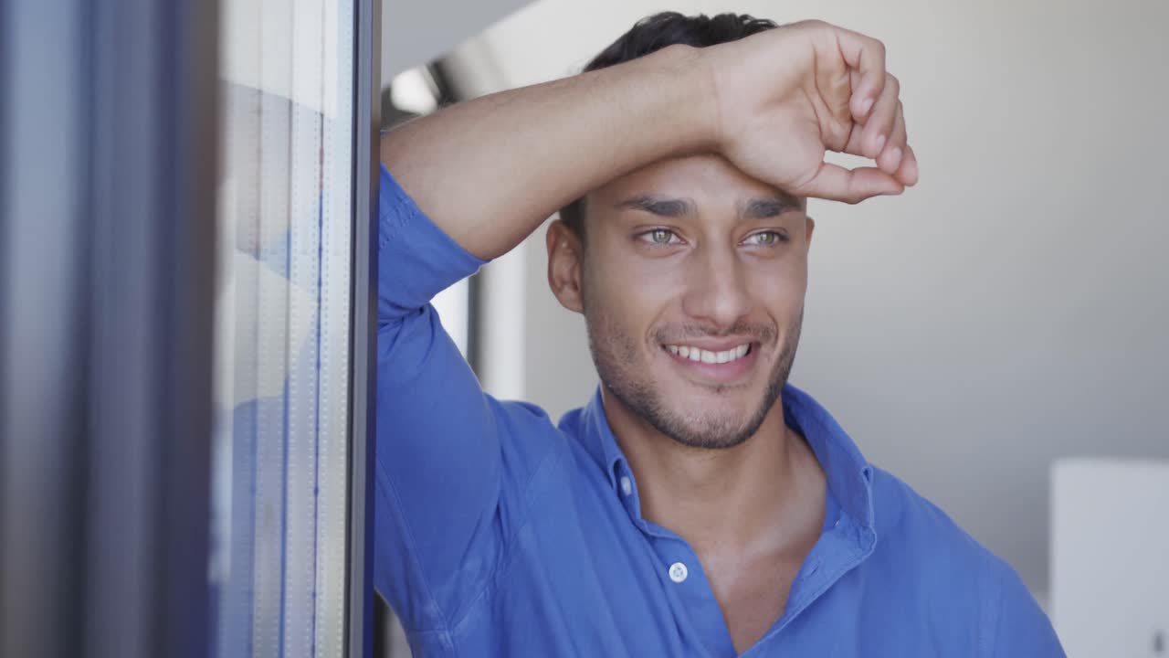 feliz hombre biracial de pie en la sala de estar, inclinado y mirando por la ventana, cámara lenta