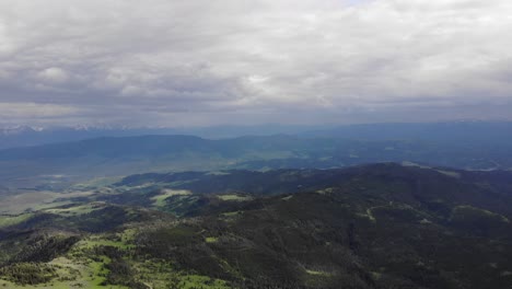 High-altitude-drone-of-green-hills,-pine-trees-and-mountains-in-the-background