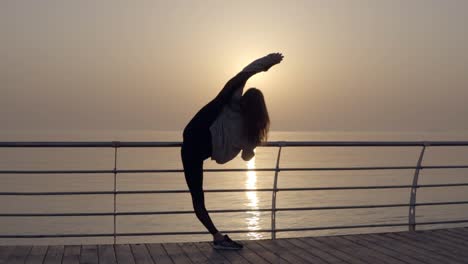 Girl-showing-incredible-flexibility-of-the-body.-She-stretches-her-legs-while-standing-on-the-promenade-near-the-sea.-Morning-sun.-Bends-into-an-arch-standing-in-a-vertical-twine