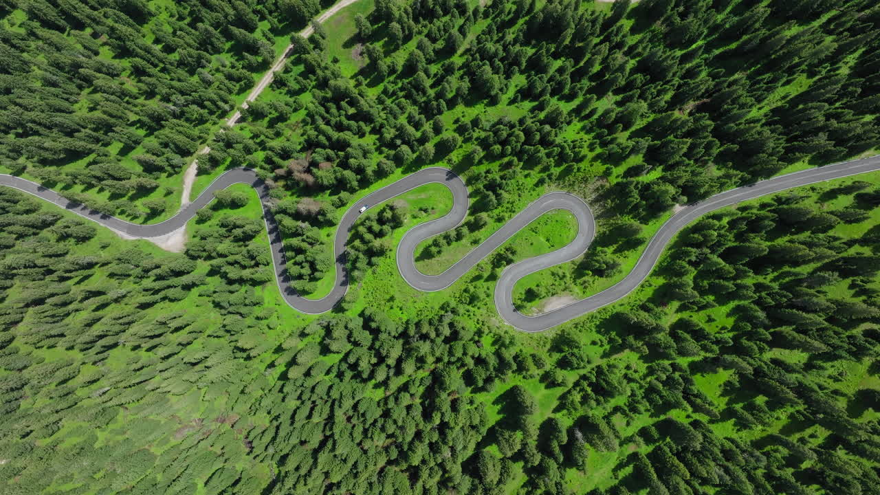 A car is driving on Strada a Serpente Passo Giau mountain pass, The snake road connecting Cortina with Colle Santa Lucia and Selva di Cadore, Dolomites, Lush green larch trees, Overhead drone shot