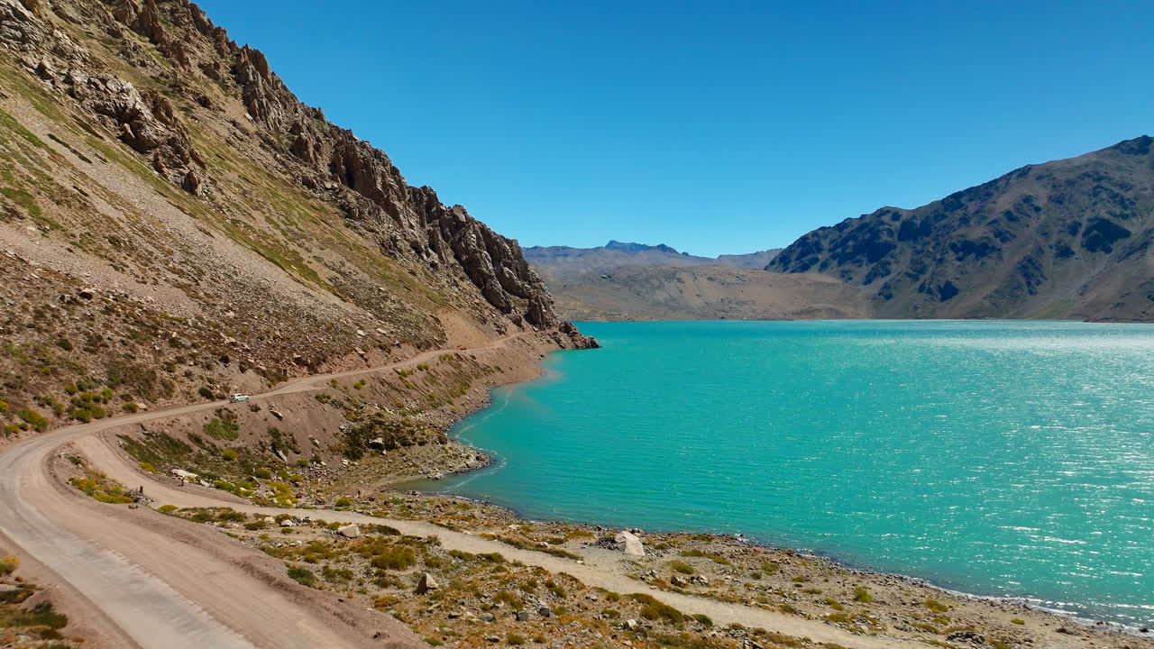 Low flight along a winding dirt road skirting a turquoise mountain lake in Cajon del Maipo, Chile, as the Andes rise on both sides of the canyon under clear summer skies