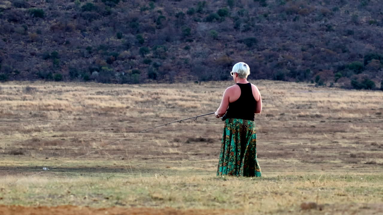 Blonde caucasian woman fishing by a lake in a flowing green dress, outdoors in nature