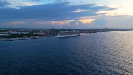 drone ascends into sky from ocean as cruise ship liner sits docked with epic sunrise in sky