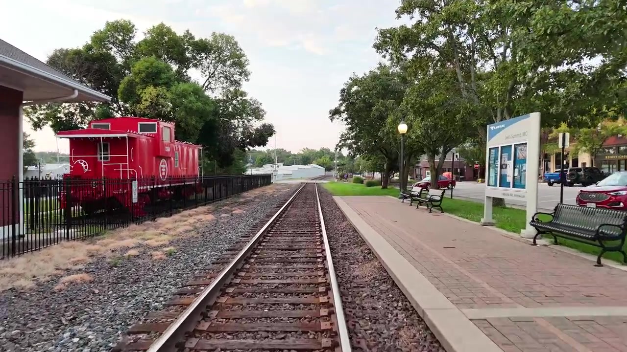 Volo di drone a bassa quota in avanti che scivola lungo i binari del treno, oltre la banchina di una piccola stazione, un vagone merci rosso, panchine della stazione e un lampione acceso sotto un cielo coperto, tenue e pallido, di prima mattina.