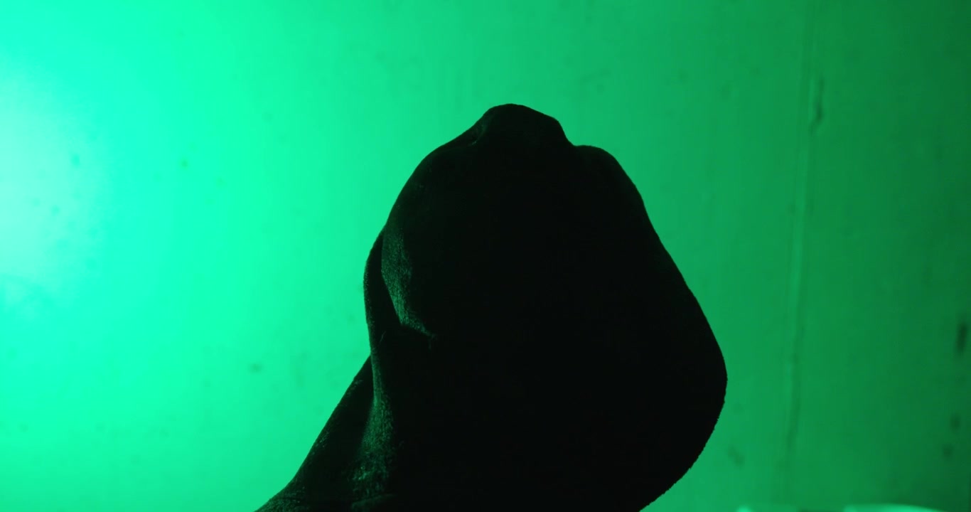 Vista posteriore di una persona incappucciata in piedi sotto un'illuminazione verde. Concetto drammatico di anonimato, segretezza, cybersecurity, mistero e identità nascosta