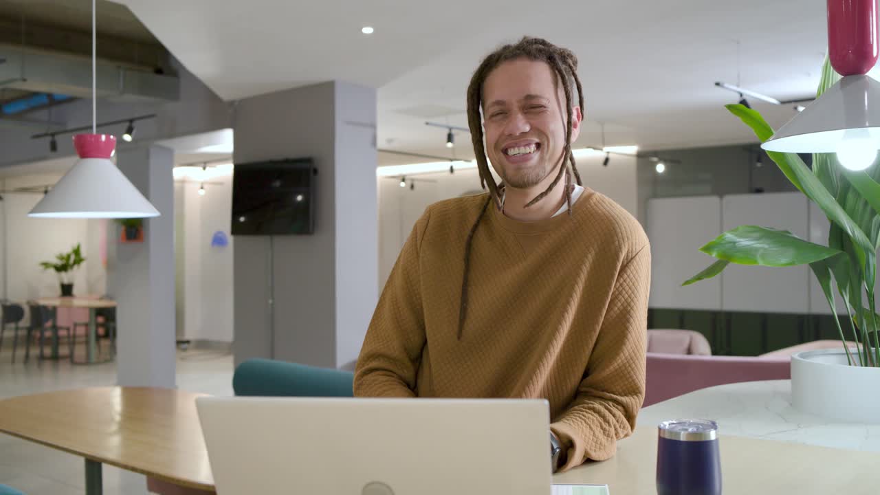 African American man laughing and smiling after camera gaze while holding phone by laptop in lounge