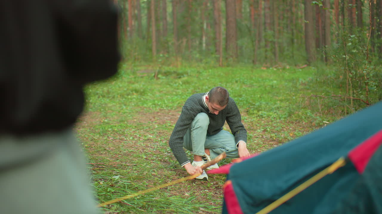 Man crouches on grass in forest hammering metal peg into ground using wooden stick while another person nearby jiggling peg, tent partially visible, surrounded by tall trees