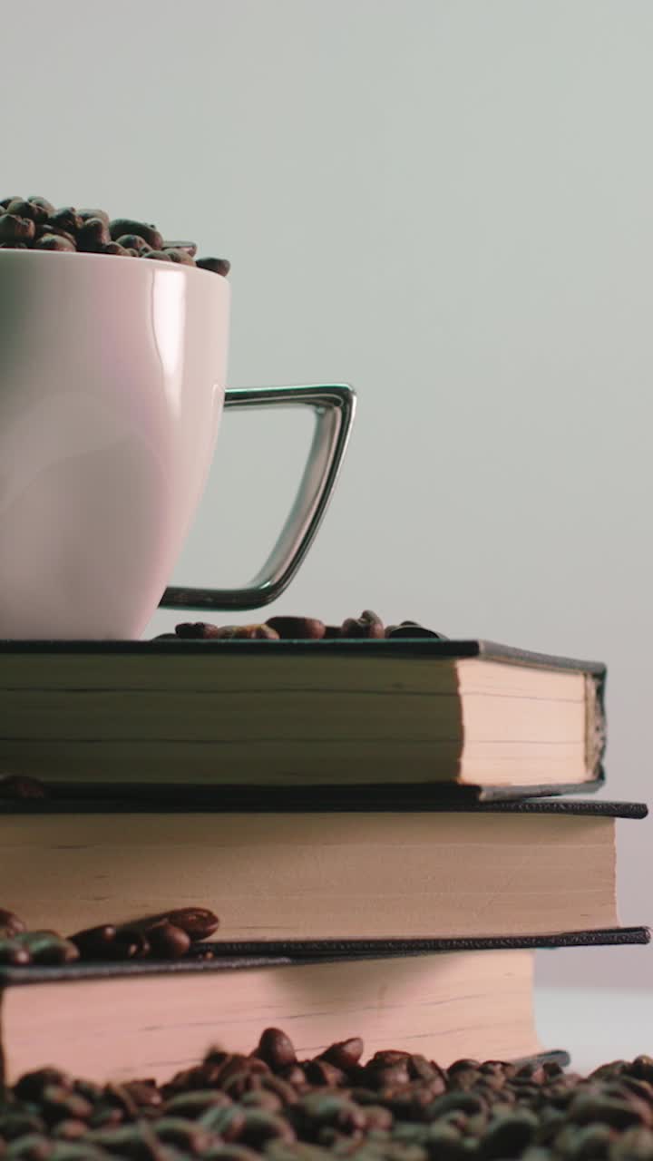 White coffee cup filled with beans rotating on stacked books with scattered beans around
