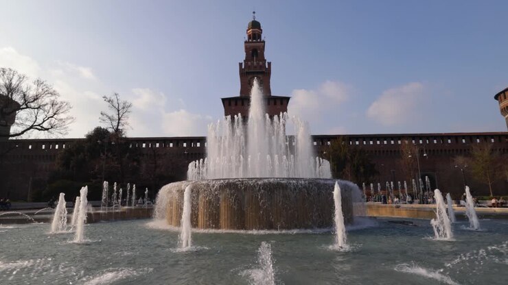 Large fountain sprays water in foreground before Sforza Castle's central Filarete Tower under clear blue sky in Milan