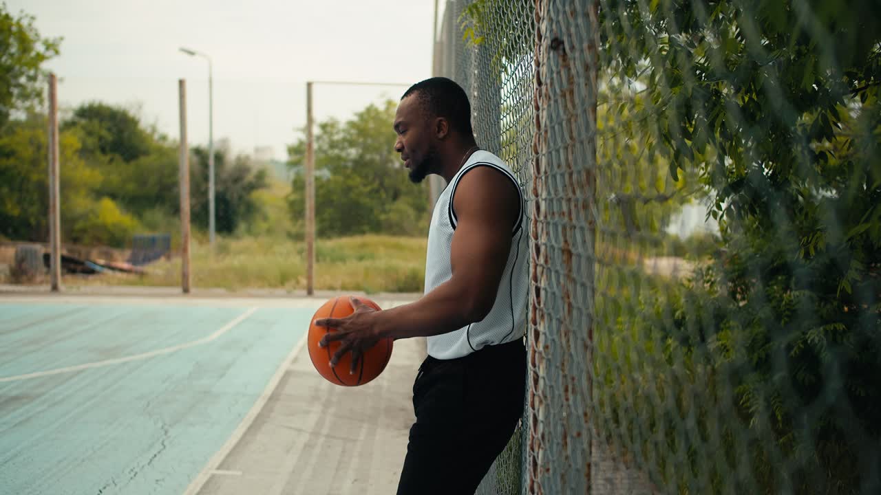 A young Black man in a white t-shirt holds a basketball in his hands and waits for his entry to the basketball court to play. Substitutes in teams of sports games