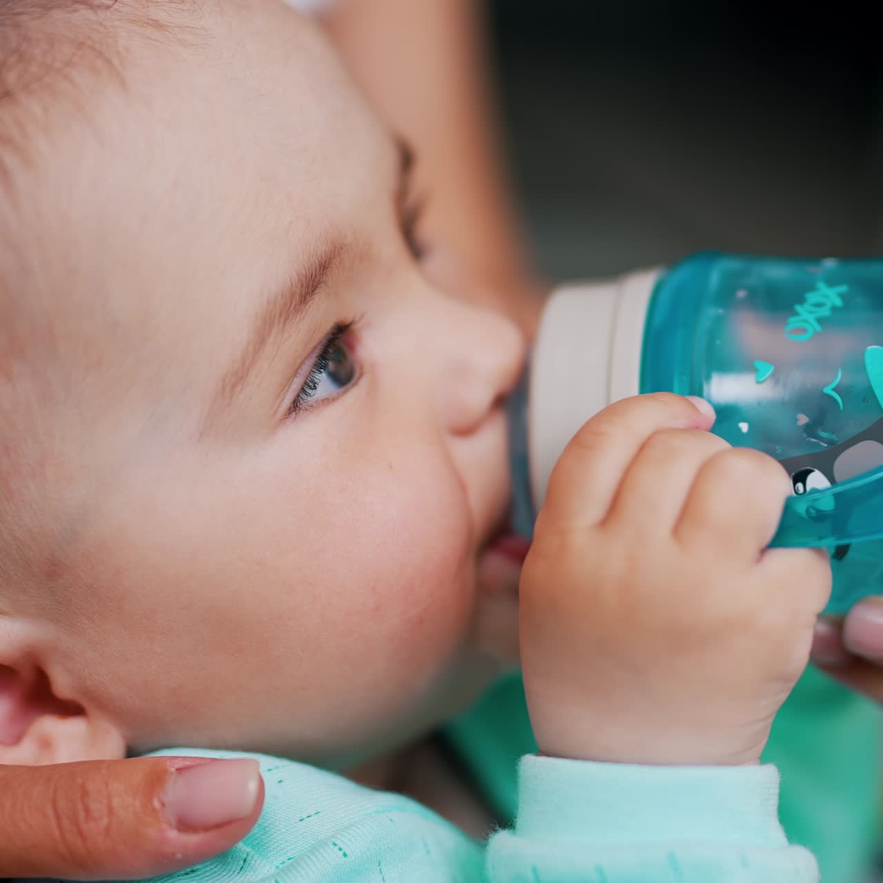 Lovely Caucasian child drinking water from a bottle and smiling. Mommy's hand hold a bottle for her son. Close up