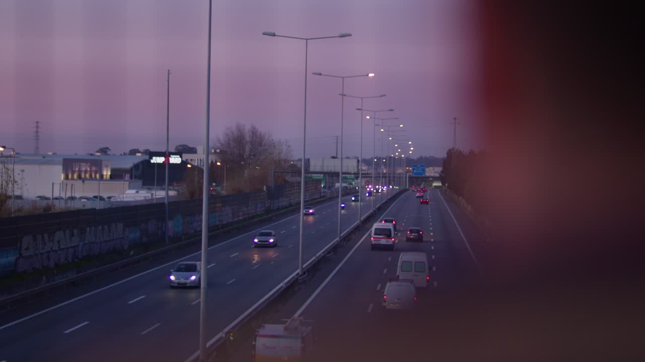 Camera reveals a highway at dusk from behind an out-of-focus man with fast cars passing by.