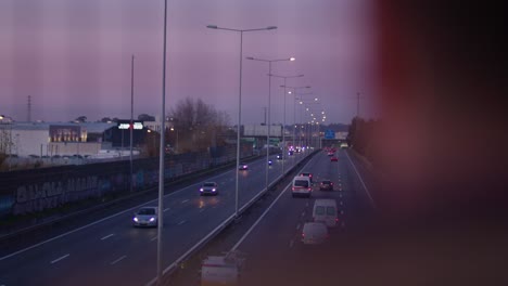 camera reveals a highway at dusk from behind an out-of-focus man with fast cars passing by.