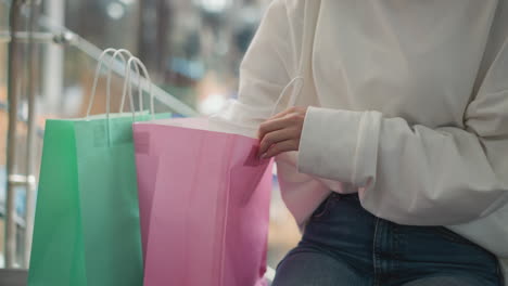 close-up of a woman in a white sweatshirt retrieving a yellow-covered book from a pink shopping bag, seated in a modern mall with glass railings and colorful shopping bags in the background