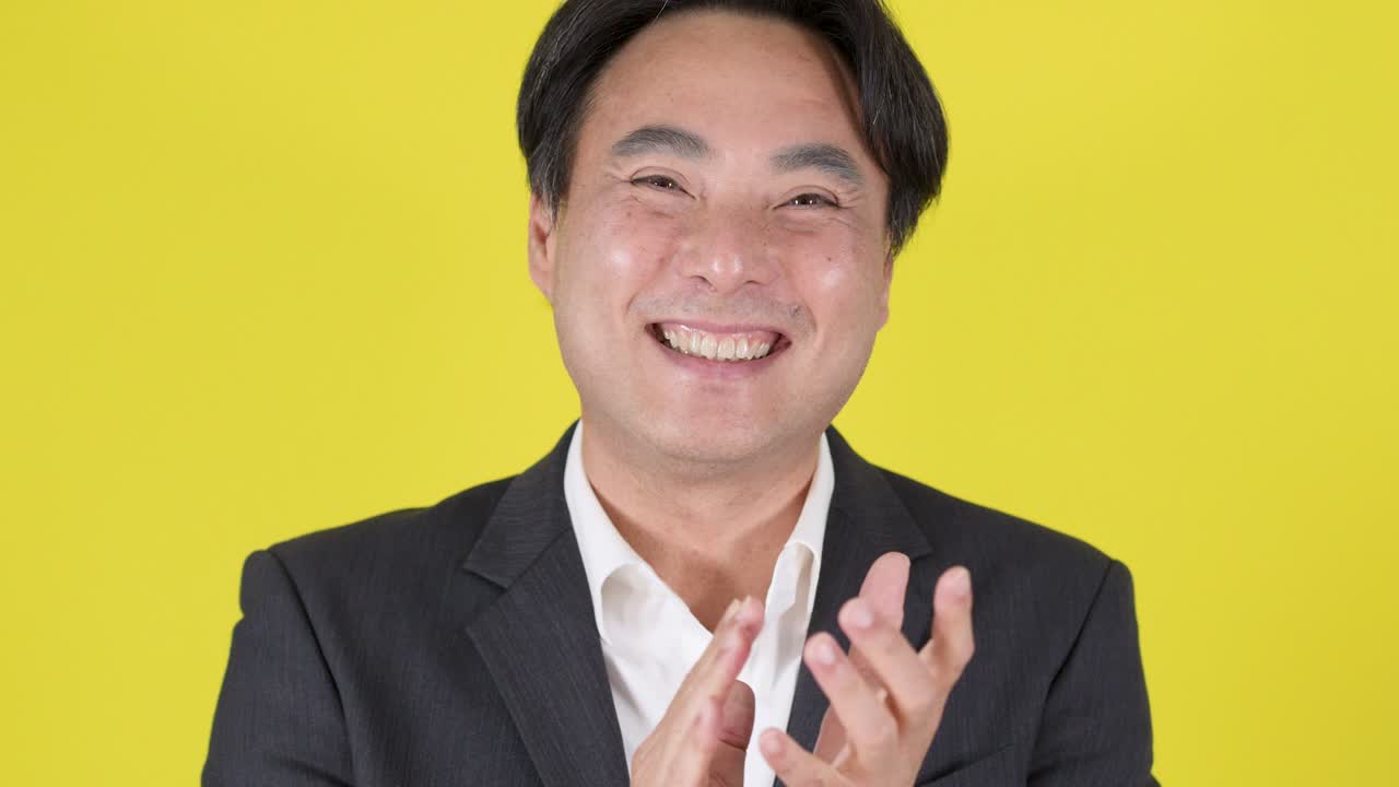 スーツ姿のアジア人ビジネスマンが、黄色の背景を前に拍手をし、笑顔で親指を立てています。