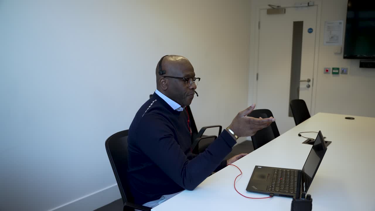 A man sits in a modern office setting, speaking and gesturing towards a laptop on a meeting table, showcasing a professional environment typically seen in business contexts