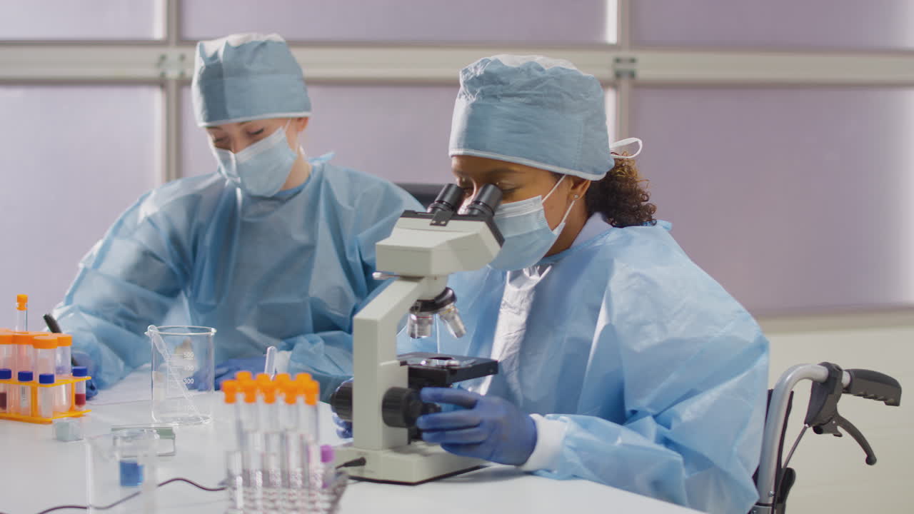 Vrouwelijke laboratoriummedewerker in een rolstoel met PBM die monsters analyseert in het laboratorium met een microscoop.