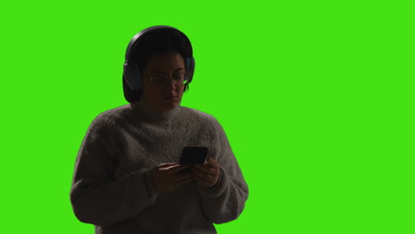 woman wearing wireless headphones streaming music from mobile phone against green screen with low key lighting 1