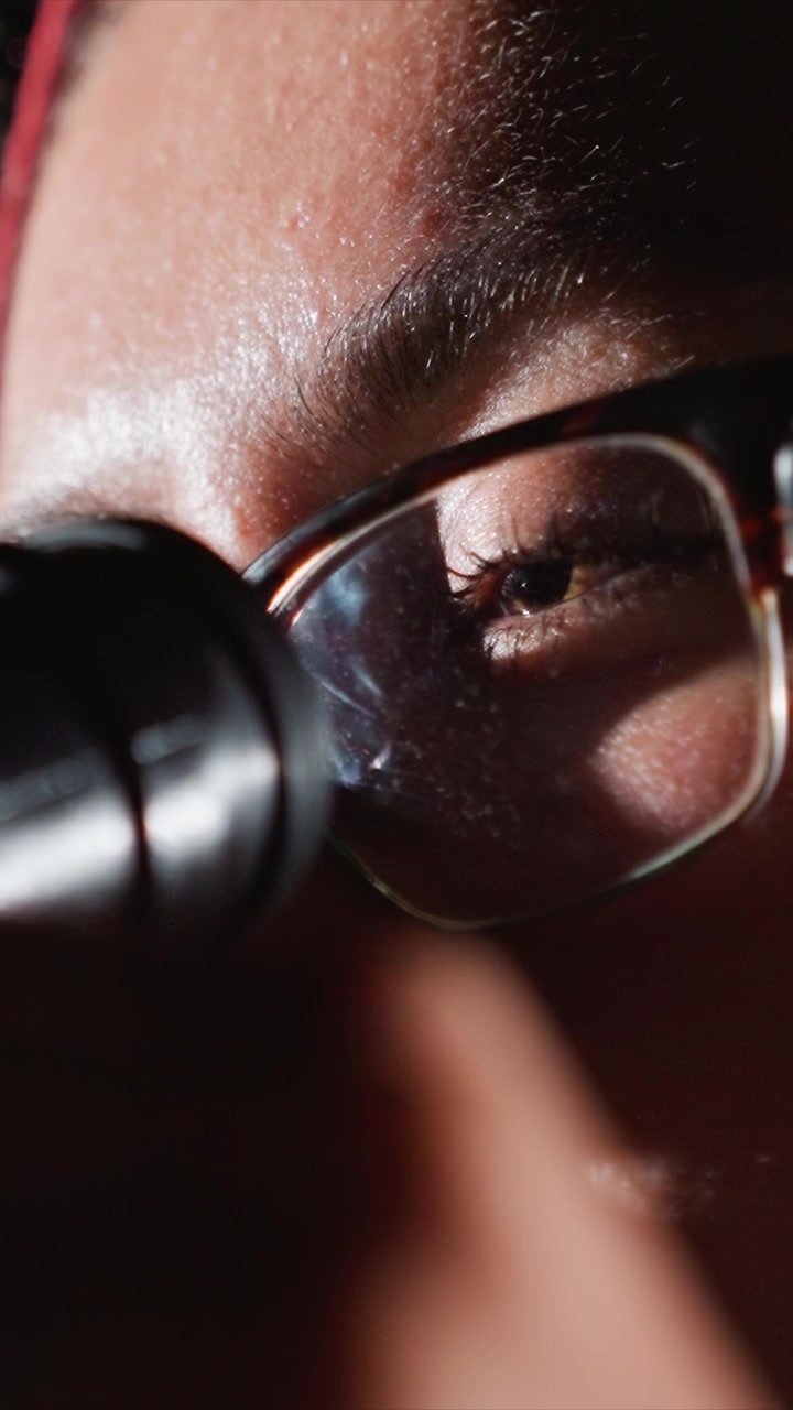 Glasses wearing individual looks through cylindrical tool. Glasses frame is clear showing side profile of face. Low camera position captures side profile from ear to nose