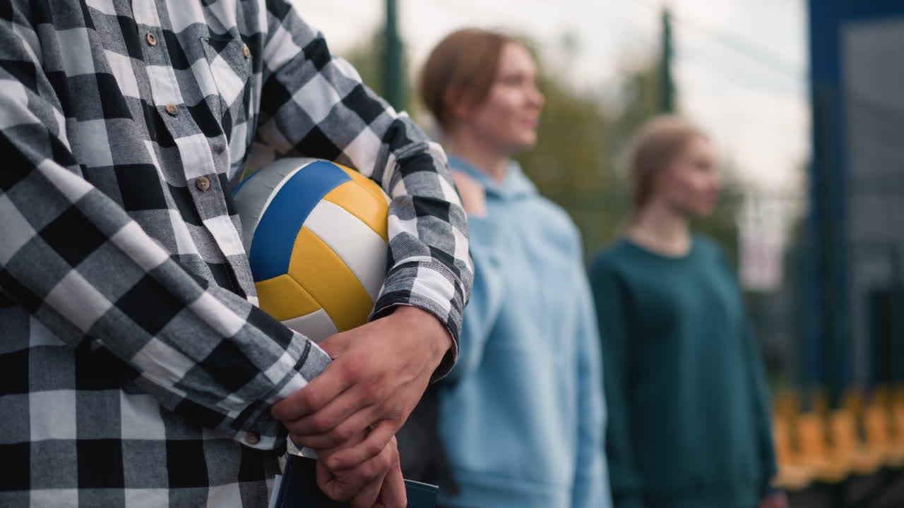 코트 세팅에서 지나가는 사람들과 초록색의 흐릿한 배경에 두 명의 웃는 여성과 함께 볼리볼을 들고 있는 체크 된 셔츠를 입은 남자의 클로즈업