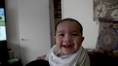 joyous smiling 4 month old indian baby looking at camera whilst sat on shoulder of adult in living room