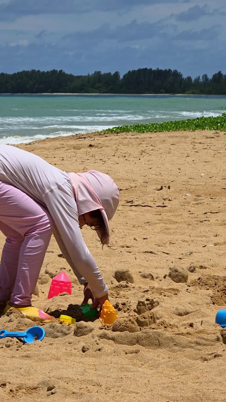 Una niña pequeña con traje de baño de cuerpo entero con protección solar juega con juguetes en la arena de una playa tropical, disfrutando de unas vacaciones de verano seguras y felices junto al mar.