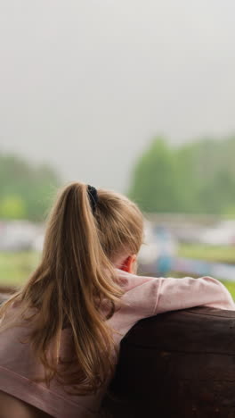 cute blonde little girl looks at modern helicopter with rotating propeller spending bored time on terrace of hotel cafe on rainy day slow motion