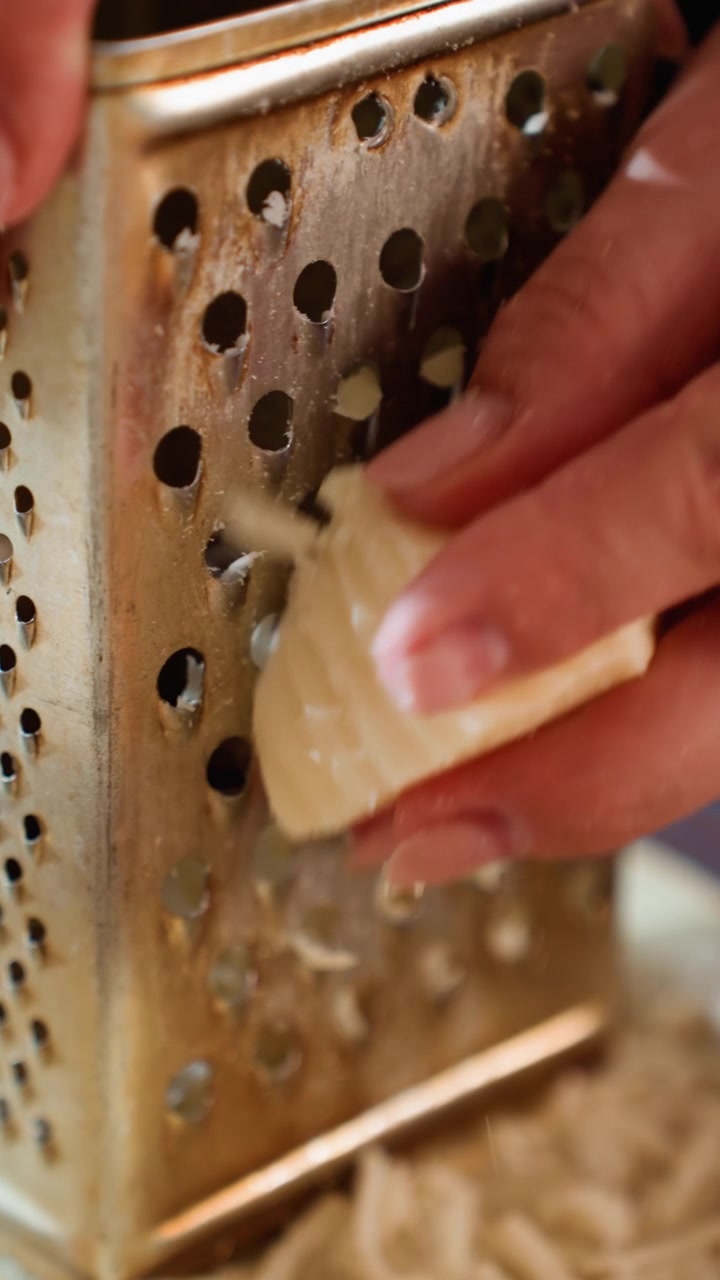 Tight side view shows hands pressing small block of warm yellow cheese across stainless steel box grater, producing thin curly shreds that fall into bowl on clean kitchen counter under soft indoor