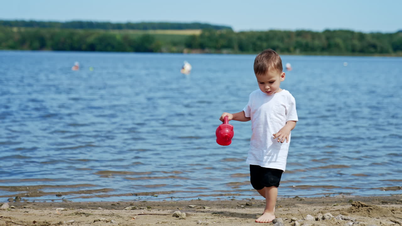 Caucasian toddler walks by the beach holding a watering can. Kid walks by the sand carefully.