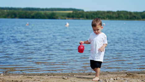 caucasian toddler walks by the beach holding a watering can. kid walks by the sand carefully.