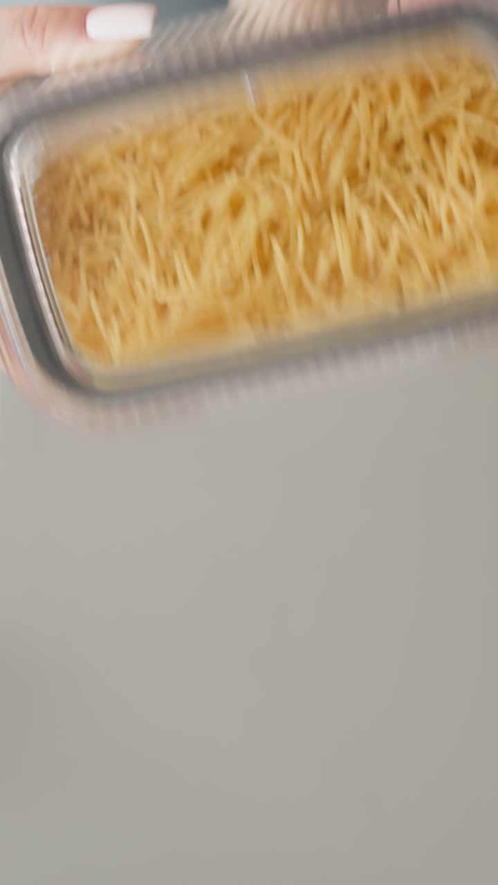 Female hand shakes closed container with spaghetti over kitchen table with serving mat slow motion. Lady packs raw food into convenient package for storage upper view