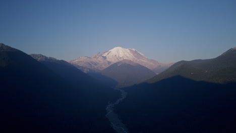 Imágenes-Aéreas-De-Las-Montañas-Alrededor-Del-Monte-Rainier-Con-Un-Valle-Con-Un-Río-En-El-Medio-Con-Una-Gran-Montaña-Cubierta-De-Nieve-En-El-Fondo