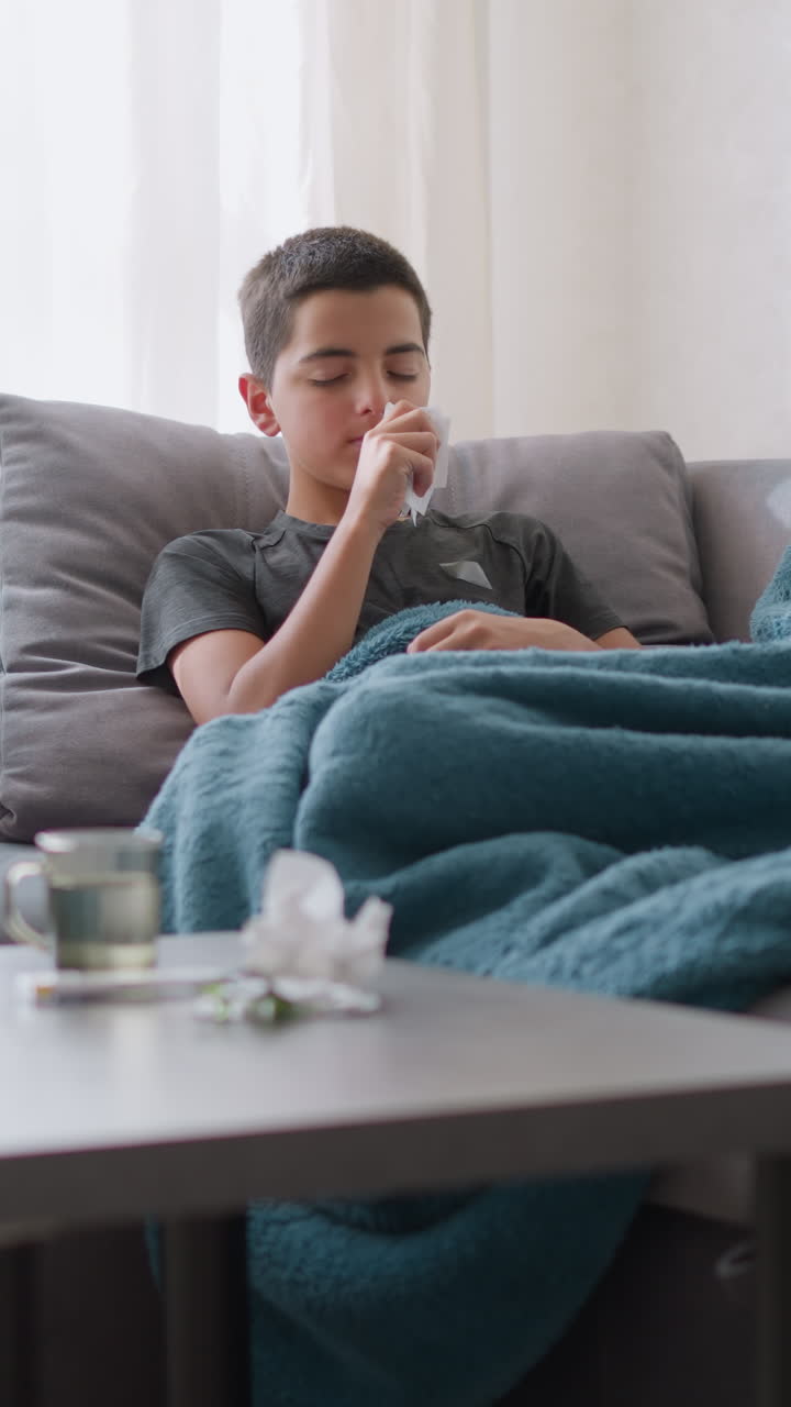 Sick little boy sitting on couch covered with blue blanket, sneezing into tissue and cleaning his nose, tissue on table with glass cup beside him. Indoor, cozy, home environment