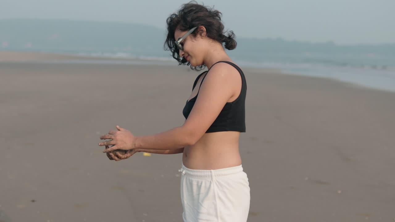 A young Asian woman with sunglasses smiles while playing with sand on the beach