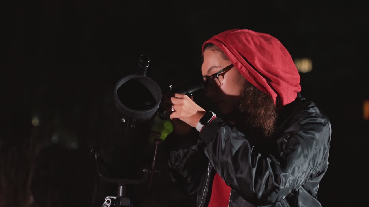 Mani bianche che regolano un telescopio su un tetto, operatore con barba, occhiali e cappuccio rosso che calibra la montatura e la messa a fuoco, skyline notturno urbano sullo sfondo, regolazione precisa dell'esposizione e del tracciamento, atmosfera di scoperta entusiasmante