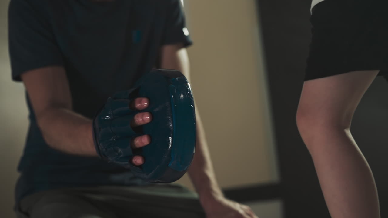 아이는 체육관에서 트레이너와 함께 무으로 패드를 때립니다. 조심스러운 강사는 무술 학교에서 작은 학생을 느린 동작으로 가르니다.