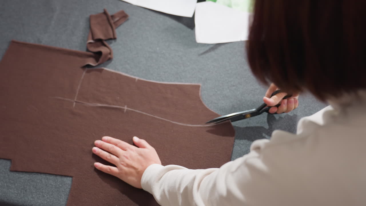 Woman placing hands on material cutting precisely along pattern lines on brown fabric using sharp scissors while creating garment pieces, surrounded by scattered paper patterns and fabrics