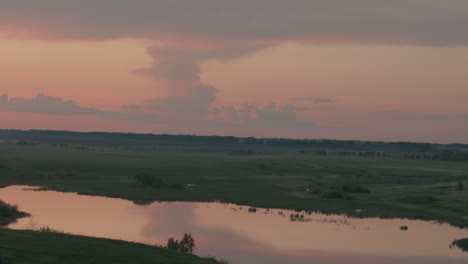scenic river winding through open prairie farmland at dusk reflecting fiery sky hues on calm water surface with distant tree silhouettes framing serene rural panorama under soft evening light