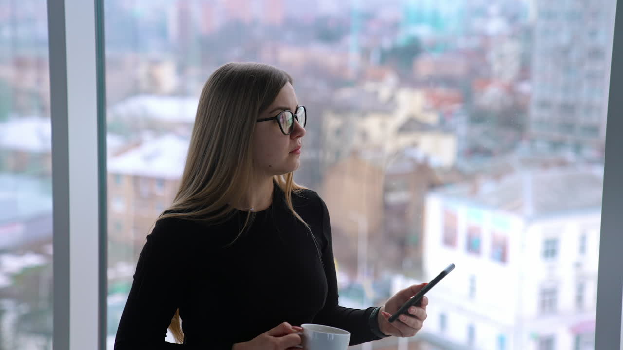 Businesswoman with a phone and a cup in office. Young woman drinking coffee and uses mobile phone on window background with a city view.