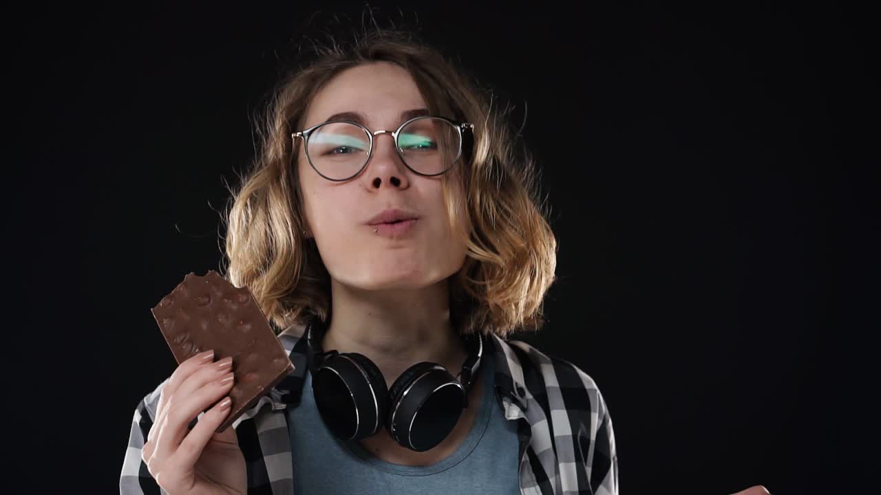 mujer joven morena con gafas y auriculares posando juguetón comer, masticar barra de chocolate con nueces aislado sobre fondo negro en el estudio. personas emociones sinceras concepto de estilo de vida