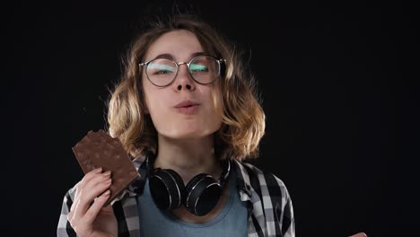 Close-up-brunette-young-woman-with-glasses-and-headphones-posing-playfully-eat,-chewing-chocolate-bar-with-nuts-isolated-over-black-background-in-studio.-People-sincere-emotions-lifestyle-concept