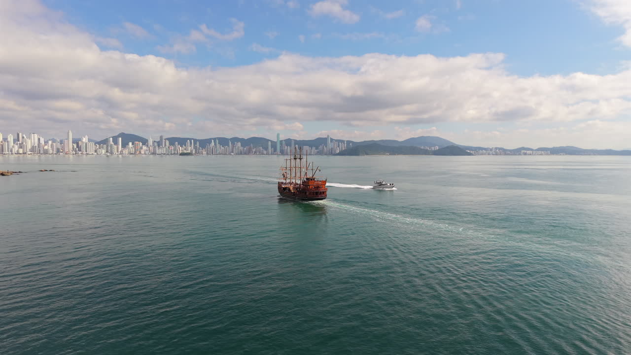 A wooden-style tourist pirate ship sails through the Gulf of Balneário Camboriú, Santa Catarina, Brazil, with the city's iconic skyline and forested Serra do Mar hills rising in the background