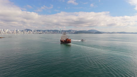 a wooden-style tourist pirate ship sails through the gulf of balneário camboriú, santa catarina, brazil, with the city's iconic skyline and forested serra do mar hills rising in the background