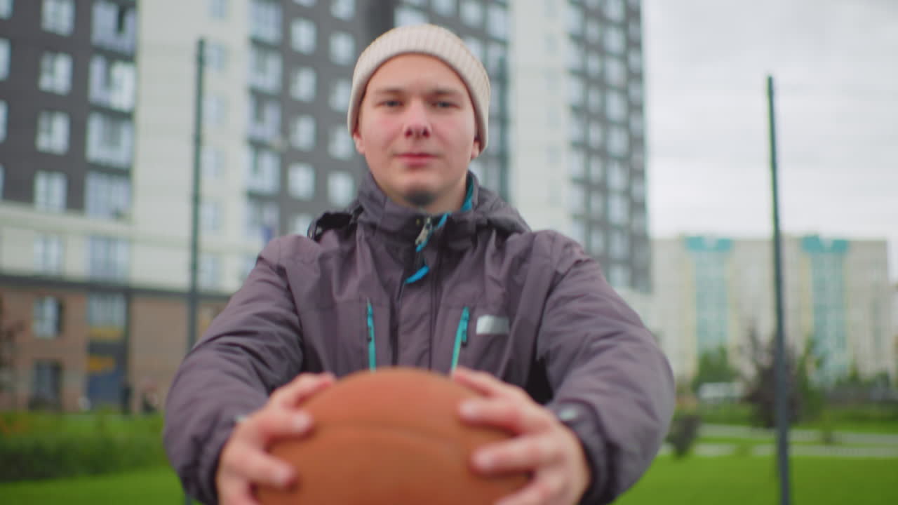 man holding basketball in park, beanie and jacket, passing ball toward camera with focused expression, residential highrises and fence in background, cloudy day atmosphere, casual outdoor training