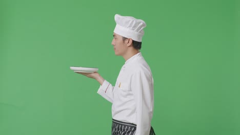 vista lateral del chef asiático sosteniendo un plato mientras camina en el estudio de fondo de la pantalla verde