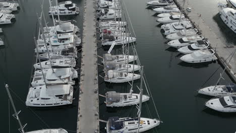 A-drone-flies-over-a-Greek-port,-capturing-the-beauty-of-white-sailboats-dotting-the-azure-sea