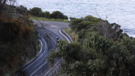 Vista-Aérea-De-Un-Motociclista-Circulando-Por-Una-Carretera-Con-Curvas-Rodeada-De-Exuberante-Follaje,-Con-El-Mar-Cerca