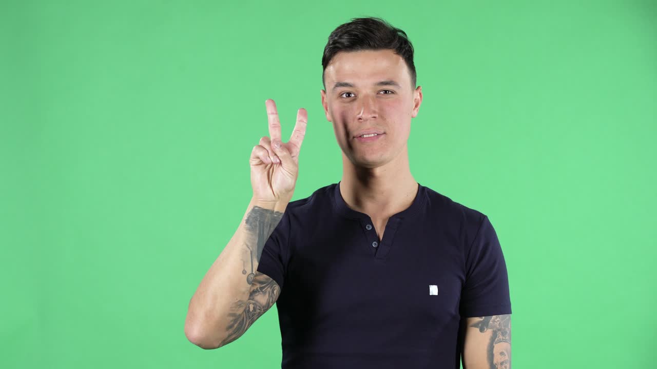 Portrait of a handsome young guy is looking at camera and showing two fingers victory gesture. Green screen
