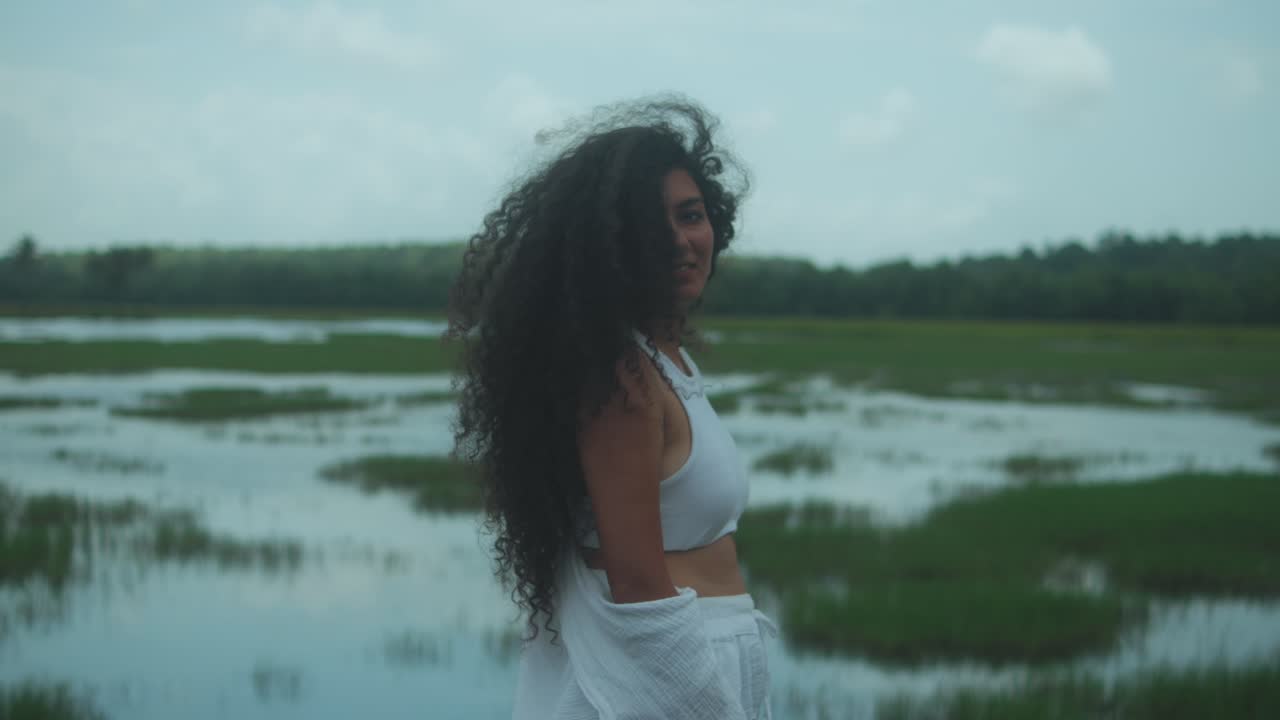 Une jeune femme aux longs cheveux noirs bouclés se tient face à des marais ou des champs inondés. Filmée de dos, elle se tourne lentement, sourit à la caméra et passe une main dans ses cheveux. Elle porte une chemise blanche
