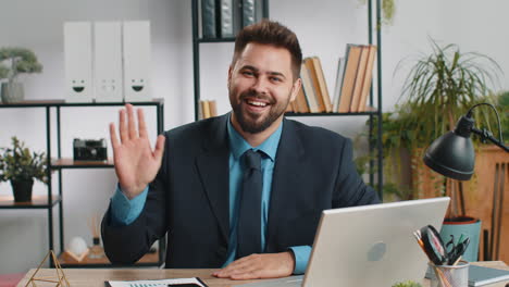 businessman working on laptop smiling friendly at camera and waving hands gesturing hello at office